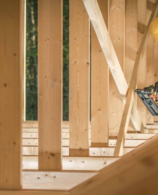 Construction Worker Contractor in His 30s With Nail Gun Attaching Wooden Elements. Wearing Hard Hat and the Wooden House Frame.